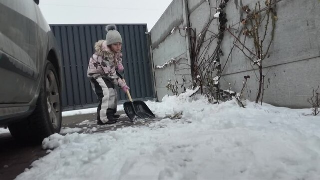 A Happy child daughter clears snow with a shovel in winter on the street near the house and near the car