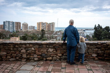 Fototapeta premium A grandfather and his grandson stand side by side, gazing at the city view from an elevated stone wall with a glimpse of the harbor in the background