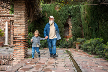 A grandfather and his grandson hold hands as they walk through a serene garden, surrounded by lush greenery and beautiful stone pathways