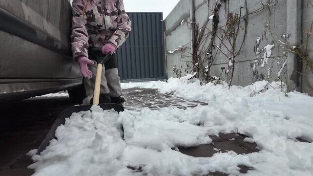 A Happy child daughter clears snow with a shovel in winter on the street near the house and near the car