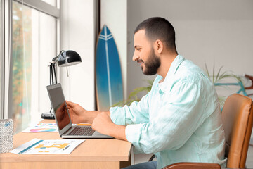 Fototapeta premium Young man sitting at workplace and using laptop for online work at home