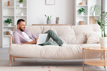 Fototapeta premium Smiling young man relaxing on sofa with laptop, remote work and online learning, cozy home environment