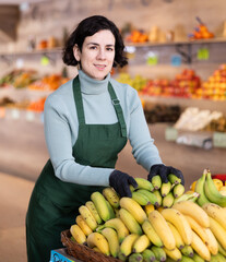 Adult female seller in uniform lays out bananas on counter in grocery store