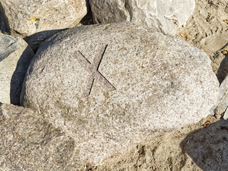 A large, smooth rock with a carved X symbol on its surface, surrounded by smaller rocks and sand.