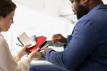 Obraz premium Woman working in dealership talking with african american man, offering color swatch samples choice. Close up of salesperson providing customer with color wheel tinting selection