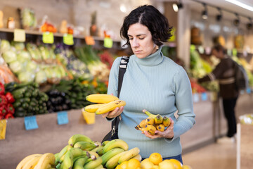 Middle-aged European woman choosing ripe bananas while standing near fruit stall