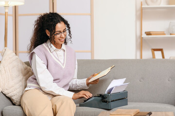 Young African-American author with typewriter sitting on sofa at home