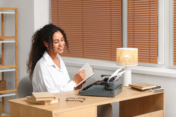 Young African-American woman using typewriter and sitting at table at home