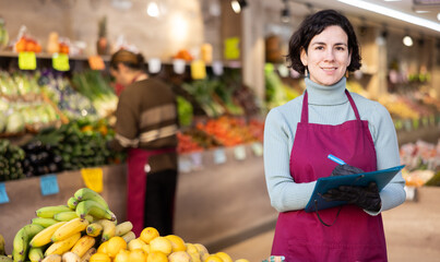 Woman store employee with documents takes inventory of goods on display case of vegetable store. Seller records quantity of goods in report, draws up document on availability of vegetables in store