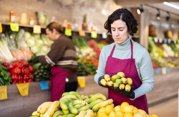 Woman shop assistant puts ripe banana fruits in box on display case, arranges assortment in vegetable shop. Worker puts vegetables in pile, in pyramid. Employee makes attractive display case