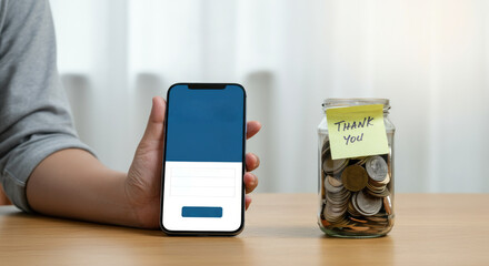 Hand holding smartphone beside jar of coins with "Thank You" note, suggesting digital payments, donations, gratitude