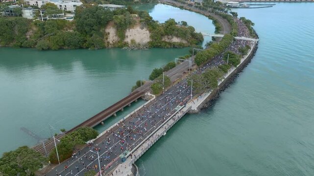 Aerial view of marathon runners on Tamaki Drive in Auckland, New Zealand. Spectators watch from a pedestrian bridge. The race promotes fitness and community engagement. PARNELL, AUCKLAND, NEW ZEALAND