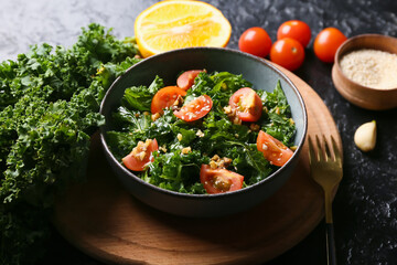 Wooden board with bowl of tasty kale salad, tomatoes and sesame seeds on dark background