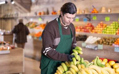 Man shop assistant puts ripe banana fruits in box on display case, arranges assortment in vegetable shop. Worker puts fruits in pile, in pyramid. Employee makes attractive display case