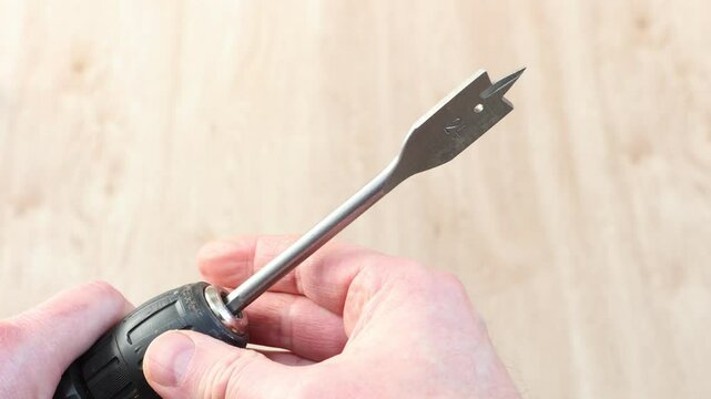 Spade bit attached to cordless screwdriver is making opening for door latch, and tip of bit appears in larger hole.