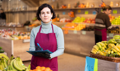 Woman store employee with documents takes inventory of goods on display case of vegetable store. Seller records quantity of goods in report, draws up document on availability of vegetables in store