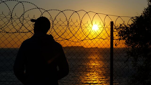 Man watching sunset behind barbed wire fence. Male silhouette wearing baseball cap standing near barbed wire fence, gazing intently at golden sunset, symbolizing freedom and contemplative solitude