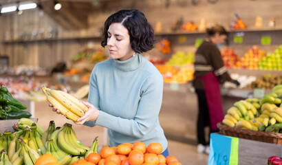 Adult woman buyer choosing bunch of fresh bananas in vegetable shop