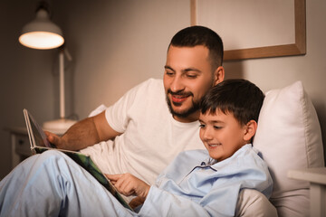 Young father and his little son reading book together in bed at home