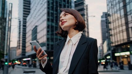 A woman in a suit checks her phone while holding a bike in a cityscape. The video uses a low-angle shot to emphasize urban modernity.