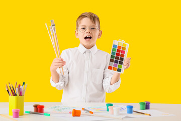 Little boy holding watercolor paints and brushes behind table with drawing supplies on yellow...