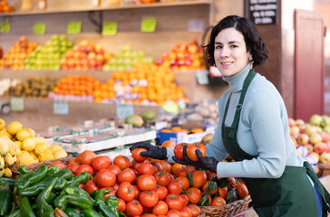 Adult woman seller in apron puts fresh tomatoes on display in vegetable shop