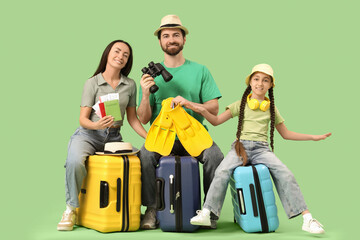 Smiling family of tourists sitting on suitcases with travel accessories on green background
