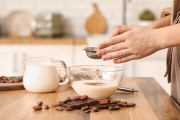 Woman adding cocoa into bowl with milk on table in kitchen. Closeup