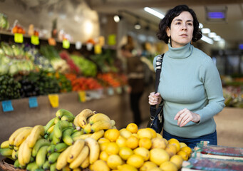 Middle-aged woman stands in a supermarket near a fruit stall and looks at the products with interest. Customer chooses fresh fruit in the grocery department