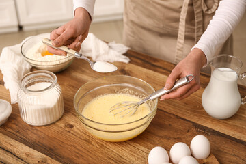 Woman adding spoon of sugar into bowl with dough at table in kitchen. Closeup