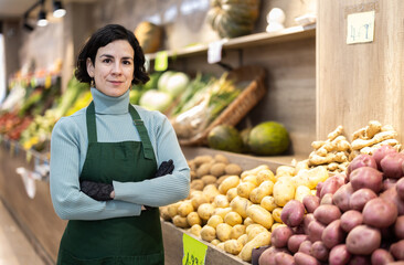 Woman store employee in apron and gloves stands in sales area of vegetable store, taking break from work. Seller has finished decorating window display, waiting for customers.