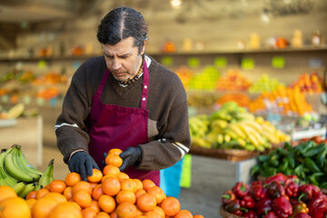 Shop seller puts goods on display case. Man creates pyramid of tangerine, presents fresh goods. Vegetable shop near house. Spacious store with long rows of display cases, customers waiting