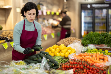 While working in shop, employee forms display case, puts ripe imported cucumber in pile. Open display of goods in self-service shop. Female employee makes attractive display case with vegetables.