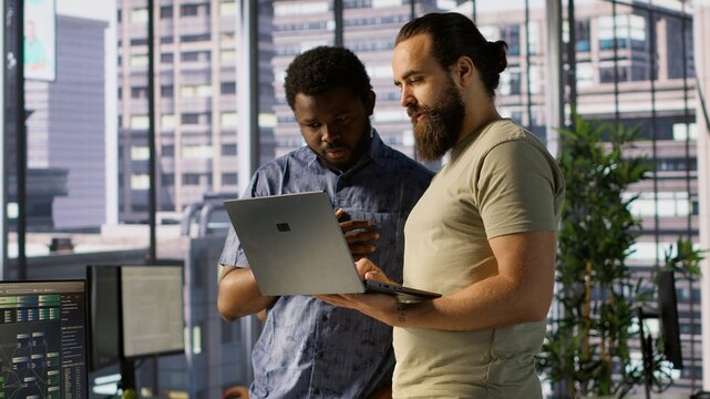 IT employees in office working together using laptop, testing and deploying programs and systems. Developer and colleague checking code on notebook, doing brainstorming in workspace, camera A