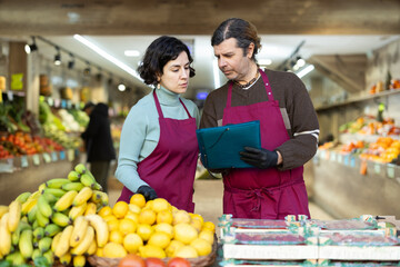 Man and woman staff in apron hold paper documents in hands, counts and records quantity of goods on display in store. Seller take inventory of goods, checks actual availability with documentary report