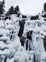 Vertical photo of large frozen waterfalls covered in snow in the French Alps. Icicles and ice formations create a winter landscape surrounded by evergreen trees.
