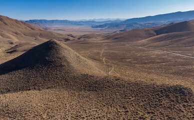 Aerial view of a remote desert landscape in California, USA. A road winds through the arid hills, connecting distant communities. Power lines run parallel to a dirt track.  Big Pine, California, USA