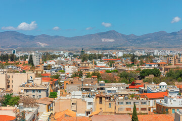 Fototapeta premium Panoramic view of Nicosia, Cyprus capital city with a mix of modern and traditional buildings, tiled roofs and greenery. Background with mountains under blue sky adding to scenic beauty of cityscape