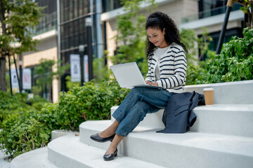 woman with curly hair sits on outdoor steps, working on laptop. She wears striped sweater and...