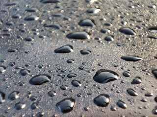 Raindrops glisten on a textured, dark surface. Subtle light reflects off the drops, highlighting the surface's grainy texture between them.