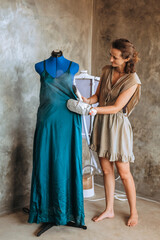 A young woman seamstress stands near a mannequin with a steamer in her hands and in a protective glove and smoothes a silk dress. A clothing designer prepares her product for a presentation. Atelier.
