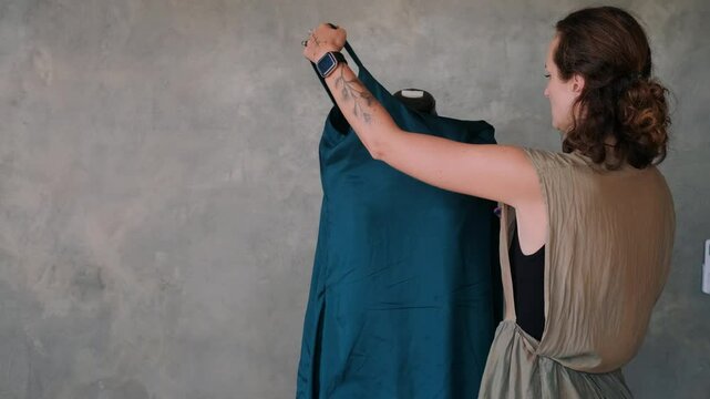 Portrait of young woman fashion designer standing next to mannequin adjusting dress, pinning it with a needle and making chalk marks to re-cut. Small studio atelier. Startup custom clothing store.