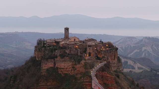 Stunning aerial footage of Civita di Bagnoregio, Italy, "dying town," perched on hilltop with illuminated streets and beautiful landscape at twilight. Aerial view