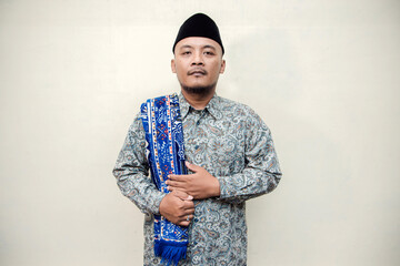 Muslim themed isolated concept photo of a tanned Asian man wearing a paisley or batik patterned shirt, black cap and prayer mat on his shoulders doing various poses on a white background.