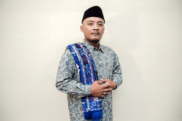 Muslim themed isolated concept photo of a tanned Asian man wearing a paisley or batik patterned shirt, black cap and prayer mat on his shoulders doing various poses on a white background.