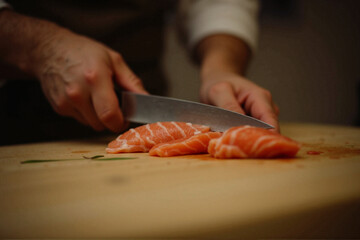 Person's hands holding a large knife and cutting a piece of salmon on a wooden cutting board. The salmon appears to be raw and has a pinkish-orange color.