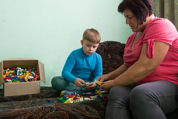 A young boy and a middle-aged woman are seated cross-legged on a couch, engrossed in playing with...