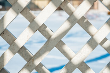Sunlight streaming through a white wooden lattice creates diamond-shaped openings, revealing a blurred, snow-covered background in a serene winter setting