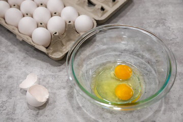 Two cracked eggs in a glass bowl view of the eggshells on the side and a carton of eggs on the background with depth of field.