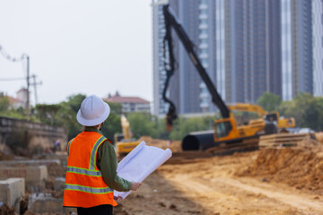 A construction engineer wearing a safety vest and hard hat holds blueprints while overseeing a...